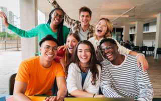 Group of diverse, smiling young adults enjoying time together, sitting around a table with a laptop.