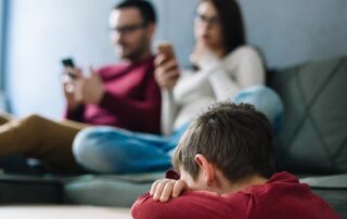 Sad child with head down while parents are distracted by their phones in the background.